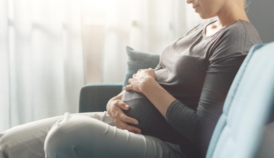 Young pregnant woman sitting on the sofa at home and relaxing, she is touching her belly and feeling happy